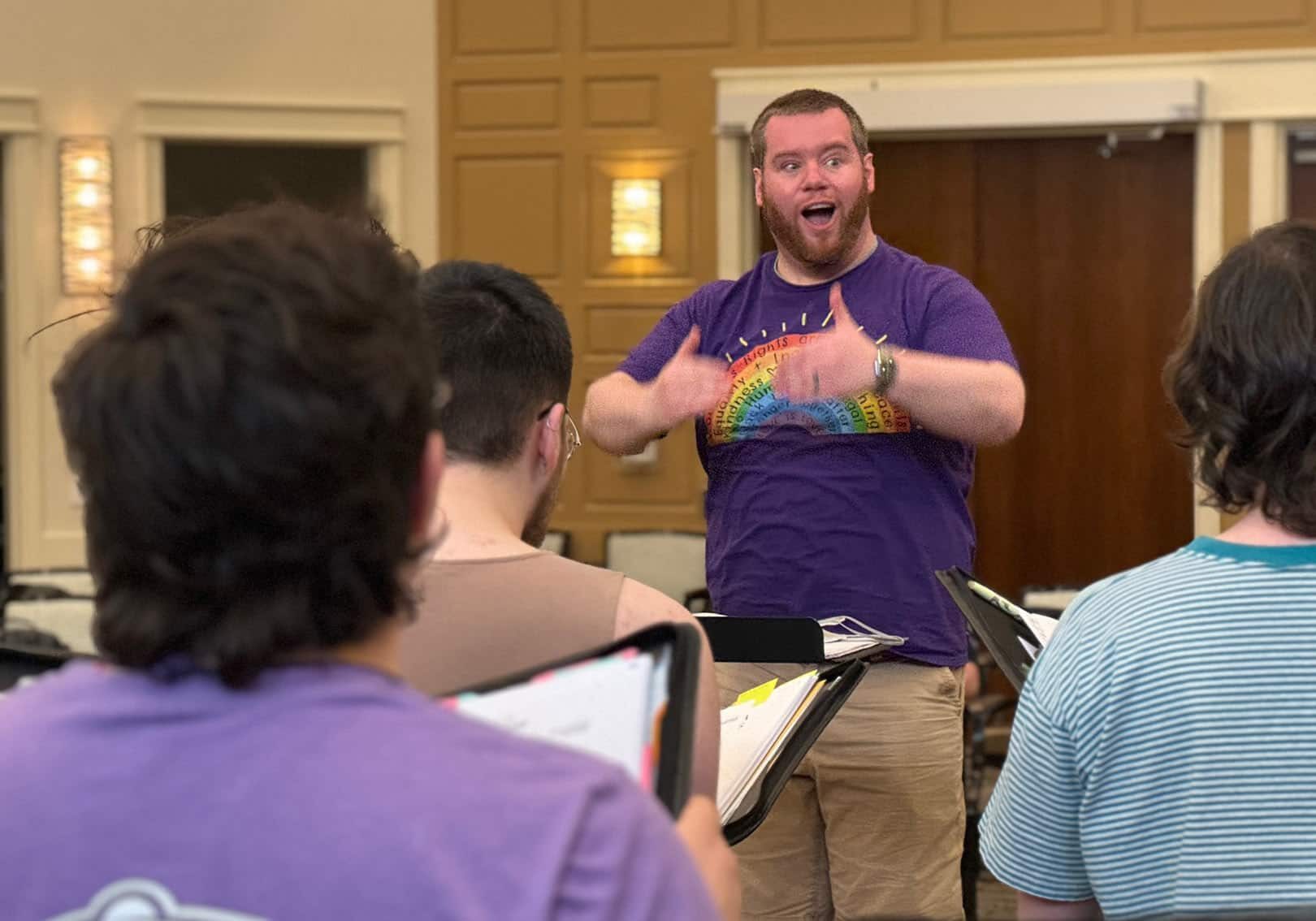 Perfect Harmony Artistic Director Kevin Hage is wearing a purple shirt with a rainbow on it while he directs the choir with expressive hands and an animated face.