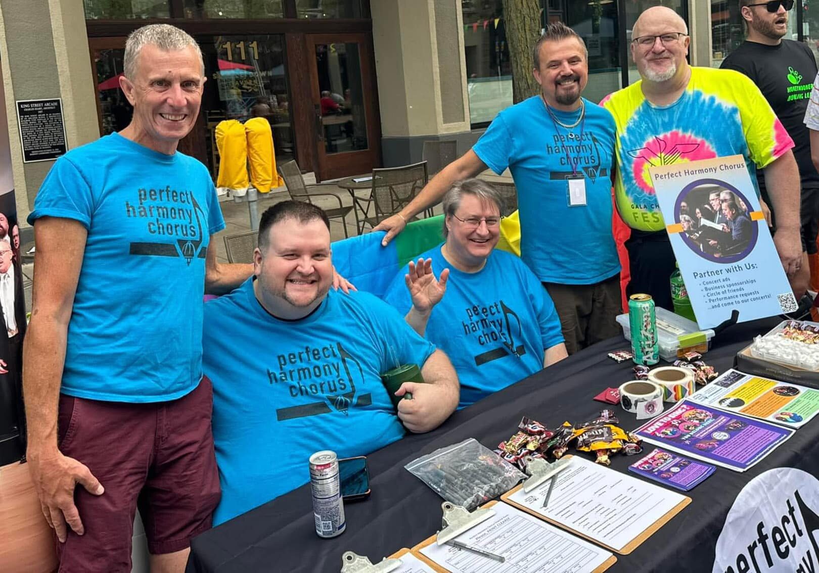 A group of Perfect Harmony singers wearing blue PHC t-shirts gather at a festival